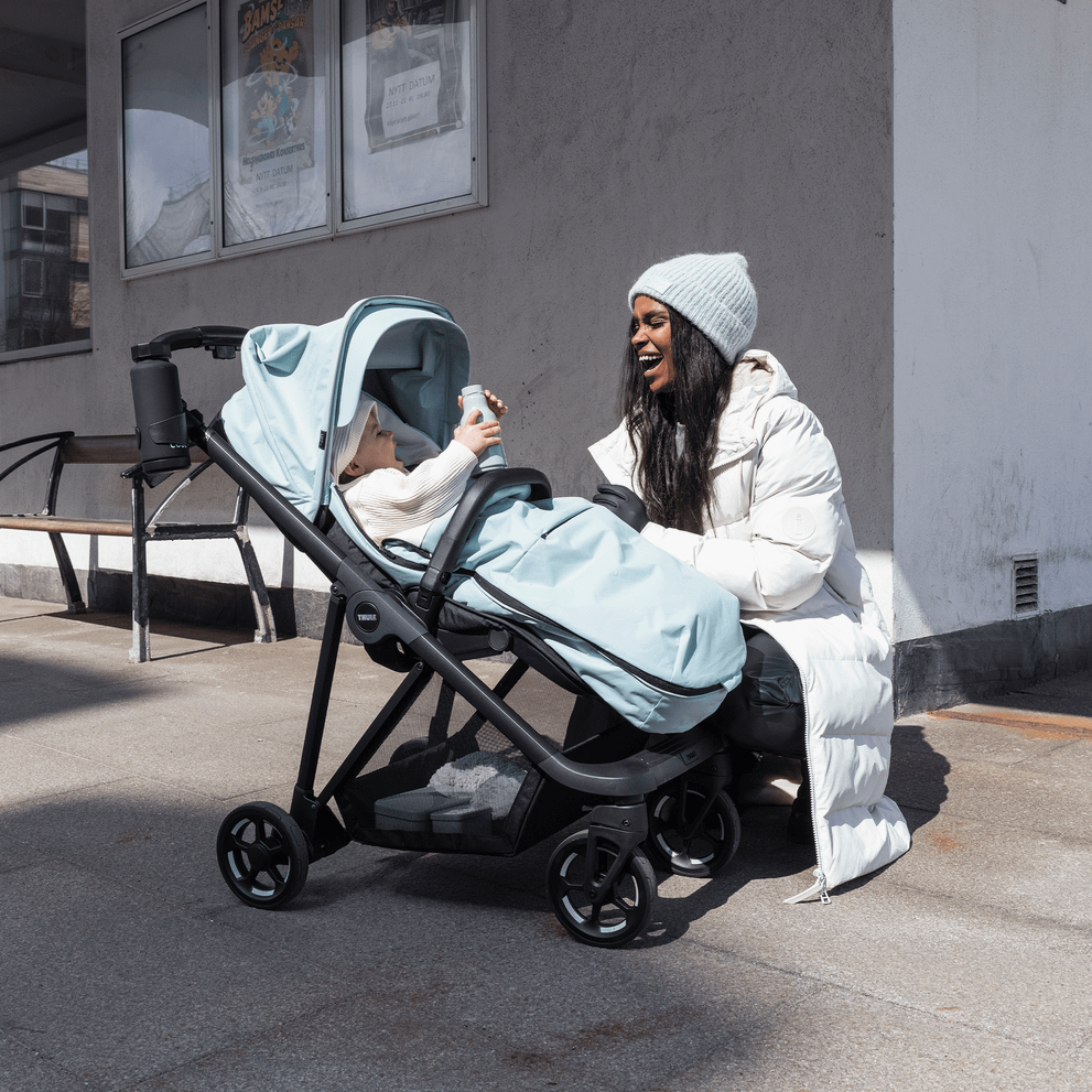 A woman in a white jacket kneels, laughing at her baby in a blue stroller with blue Thule Stroller Footmuff.