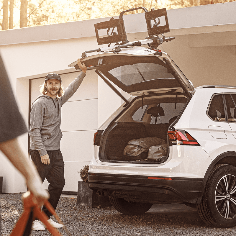 A man loads items into the open trunk that also has a Thule OutWay 2bike Platform trunk bike rack installed.