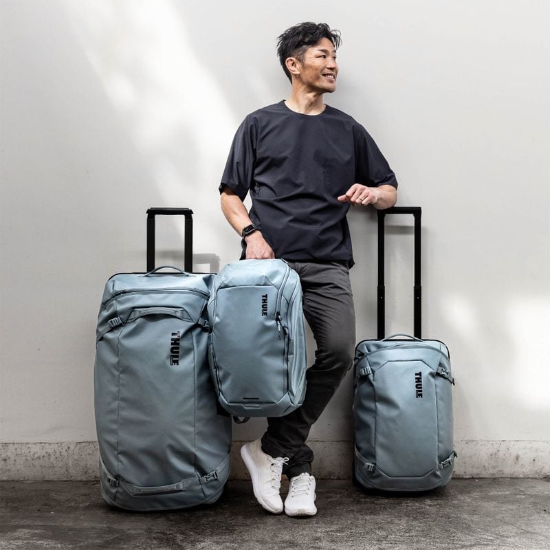 A man is leaning against a white wall with blue Thule Chasm suitcases and a Thule chasm backpack.