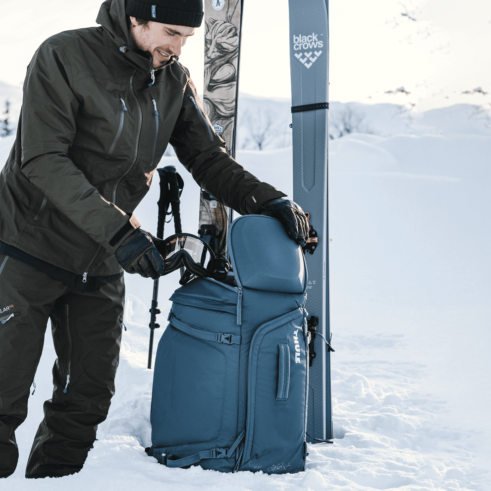 A man with skis standing in the snow loads his goggles into a Thule RoundTrip 60L Ski Backpack.
