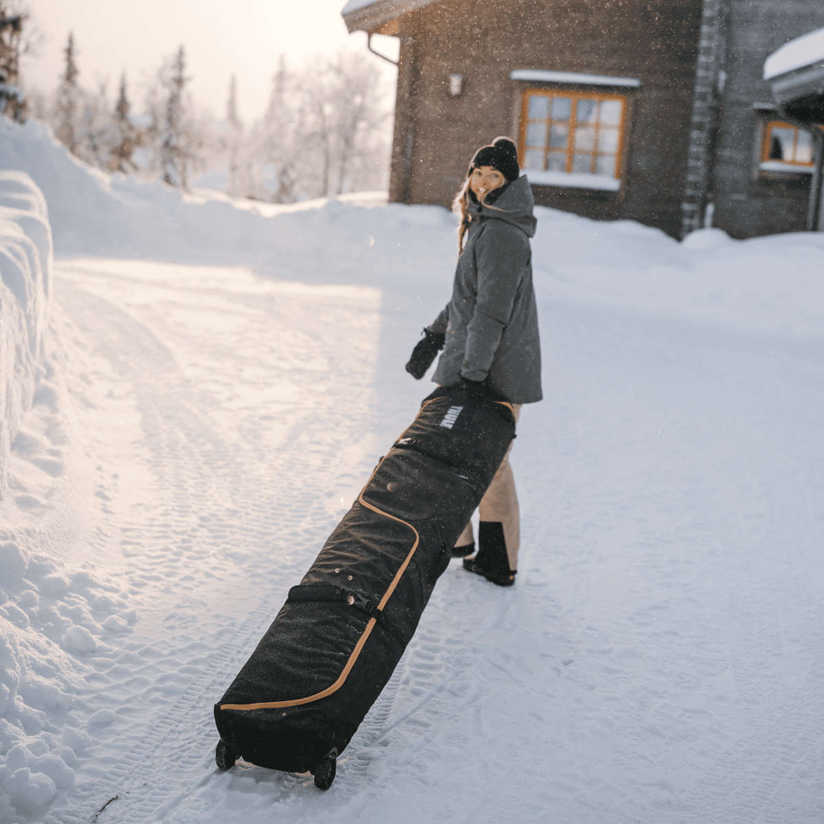 A woman stands in the snow outside a cottage wheeling her Thule RoundTrip travel ski bag.