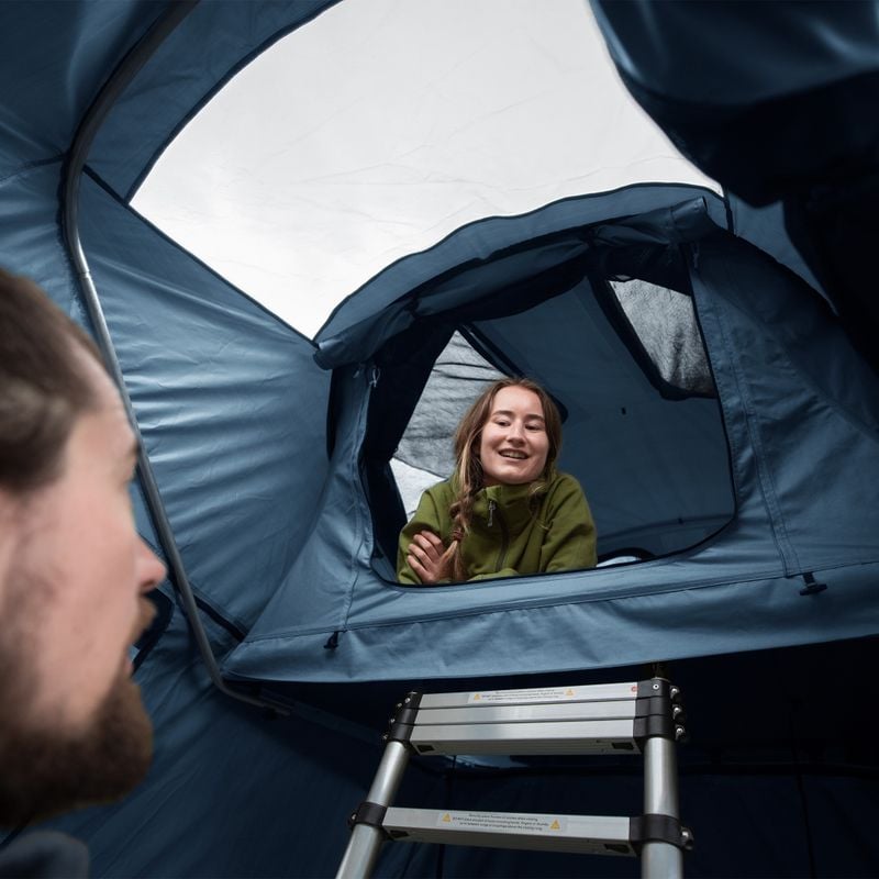 A woman looks out of a roof top tent and greets a bearded man.