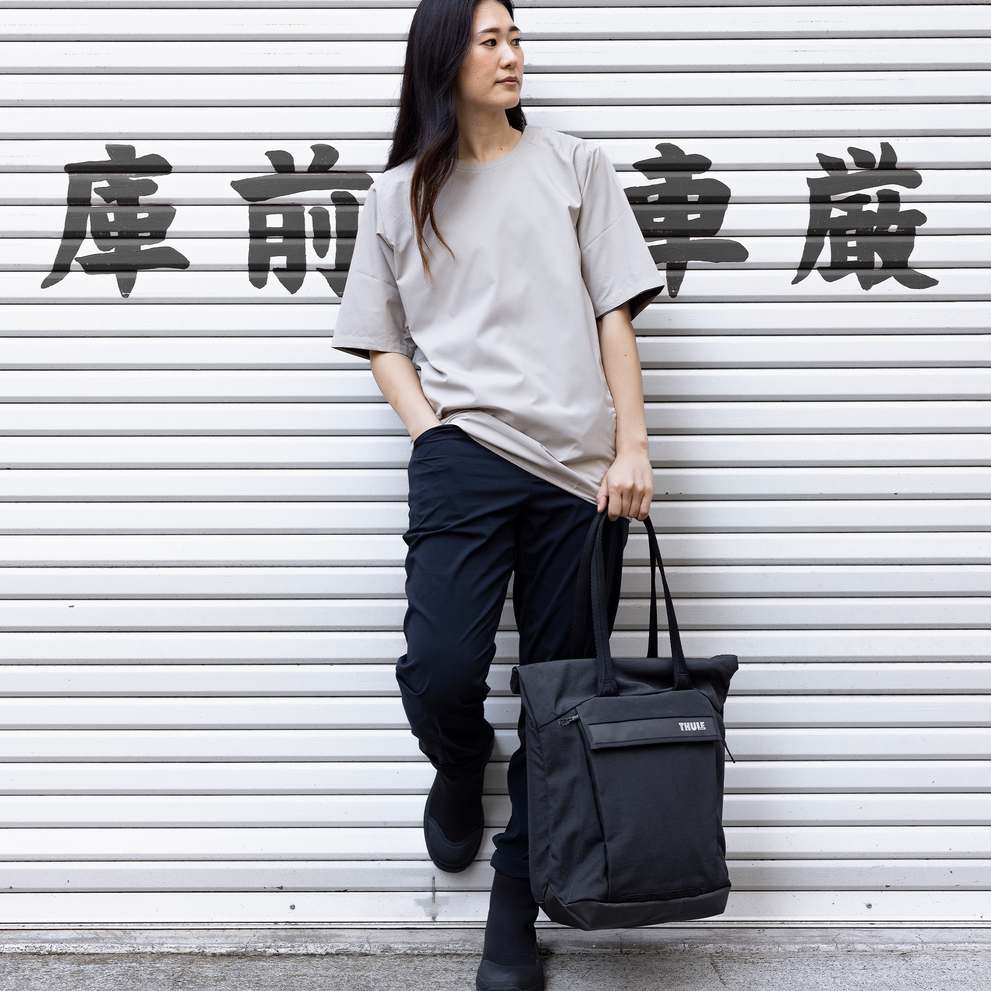 Woman stands against white wall with a Thule Paramount tote bag.