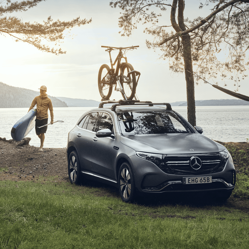 A man walks next to the lake with a SUP next to his car with a Thule UpRide roof bike rack.