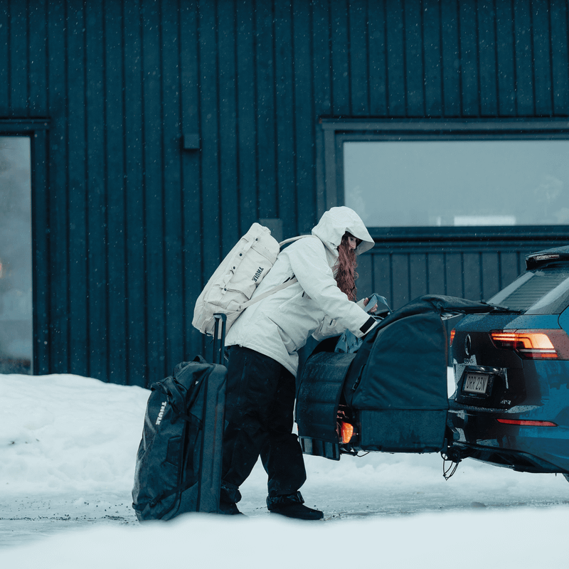 A person in a winter coat loads a large bag into the Thule Onto 2, a towbar-mounted cargo bag, attached to the rear of a blue station wagon in a snowy environment.