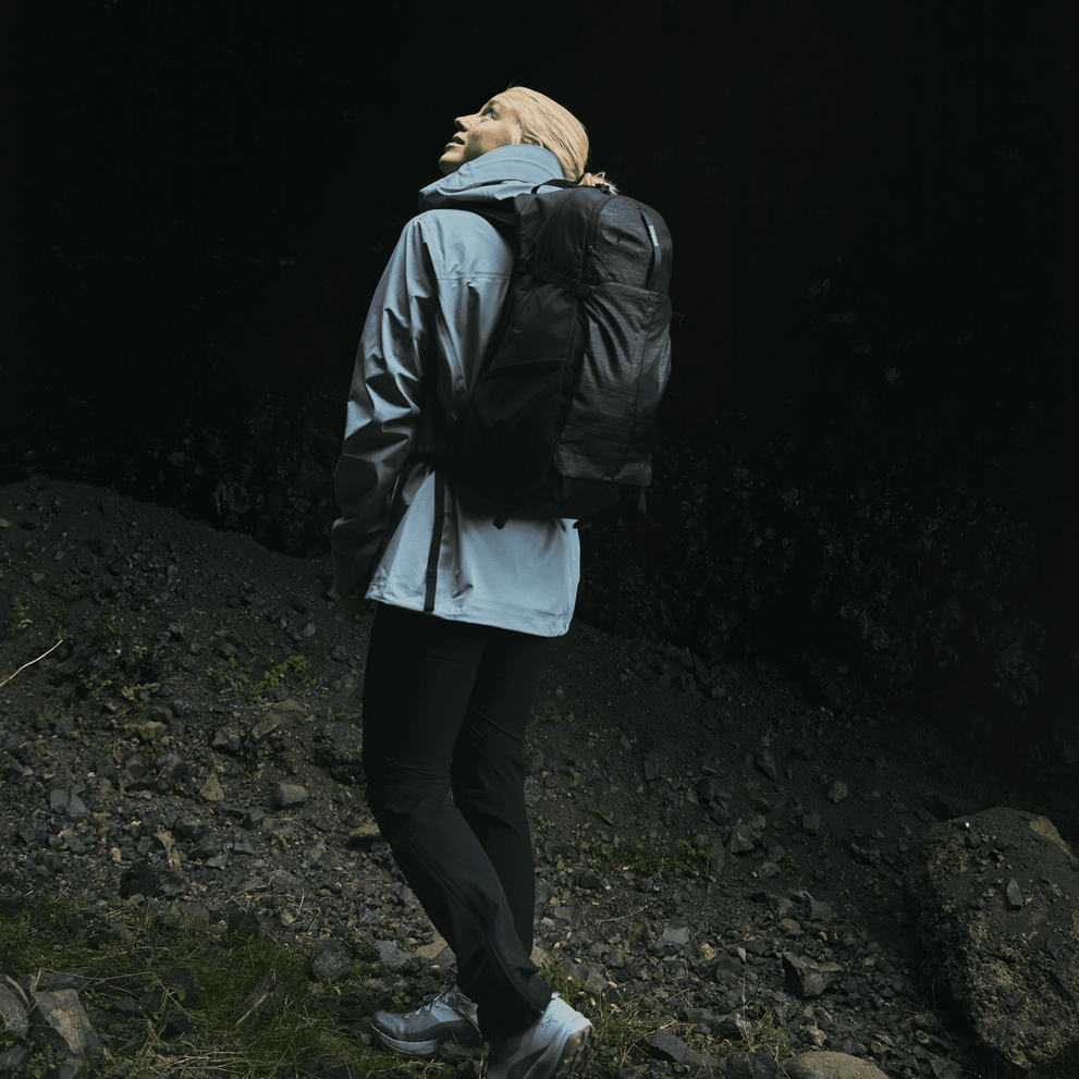 A woman in a cave looks up, wearing a blue jacket and carrying a black Thule Nanum 25L  hiking backpack.