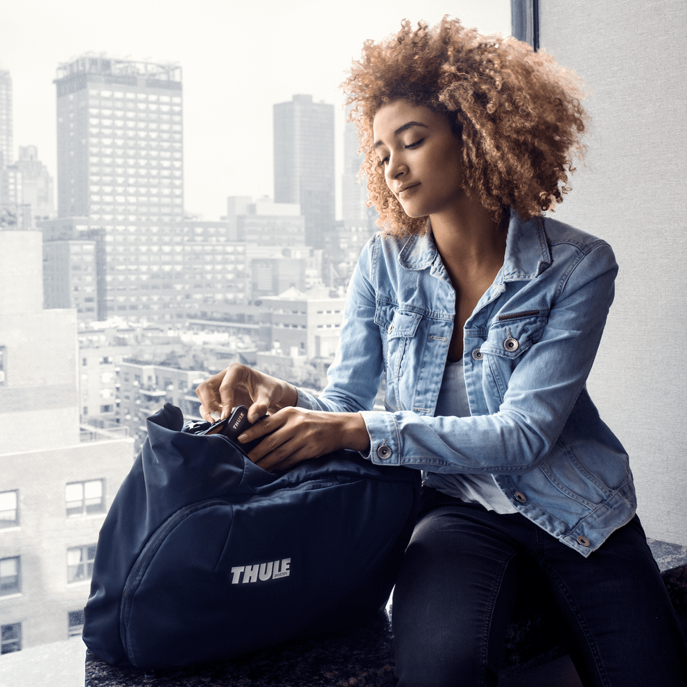 Overlooking a city landscape, a woman takes out her items from a blue Thule Subterra Travel Backpack.