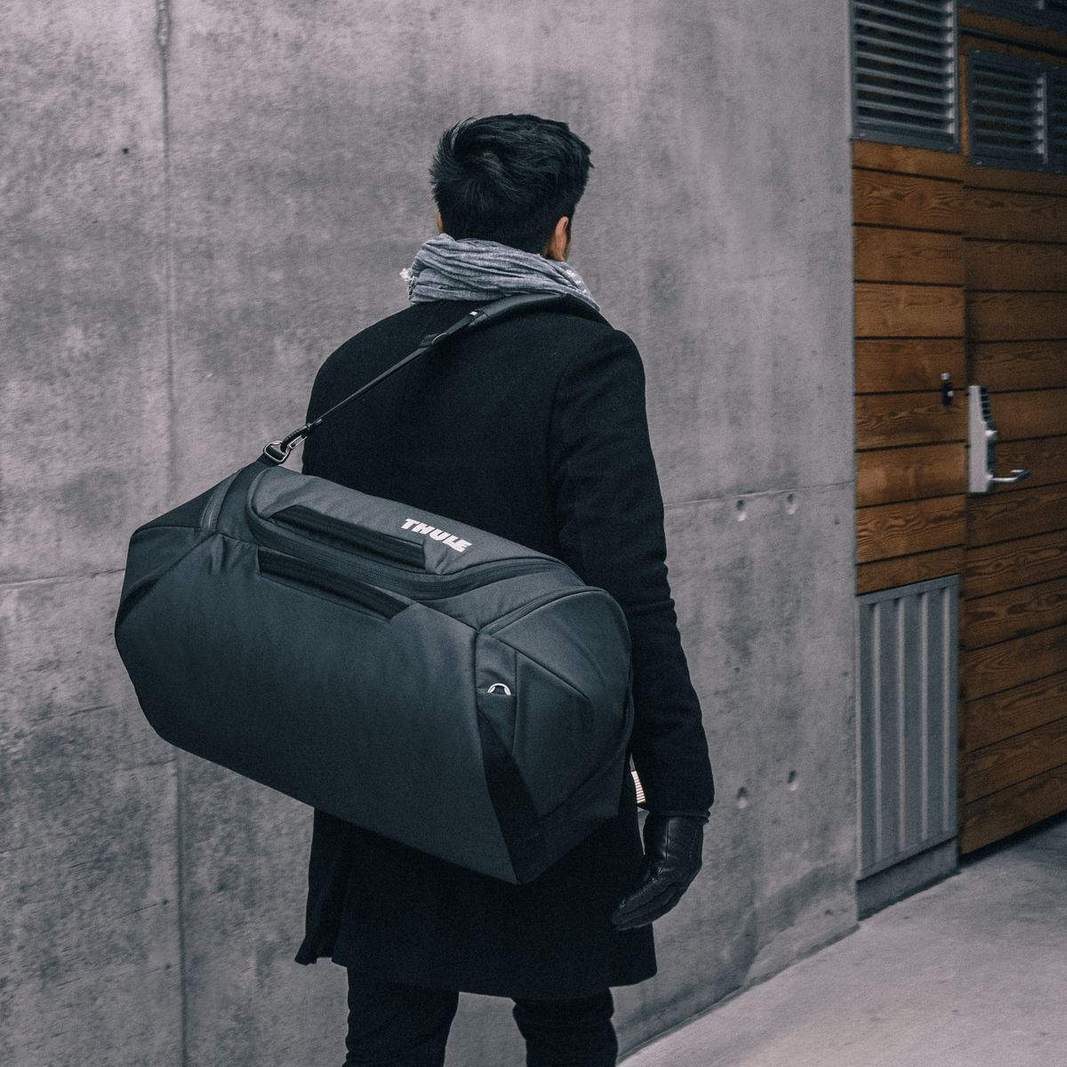 A man walks by a concrete wall holding a black Thule Subterra Weekender Duffel bag around his shoulder.