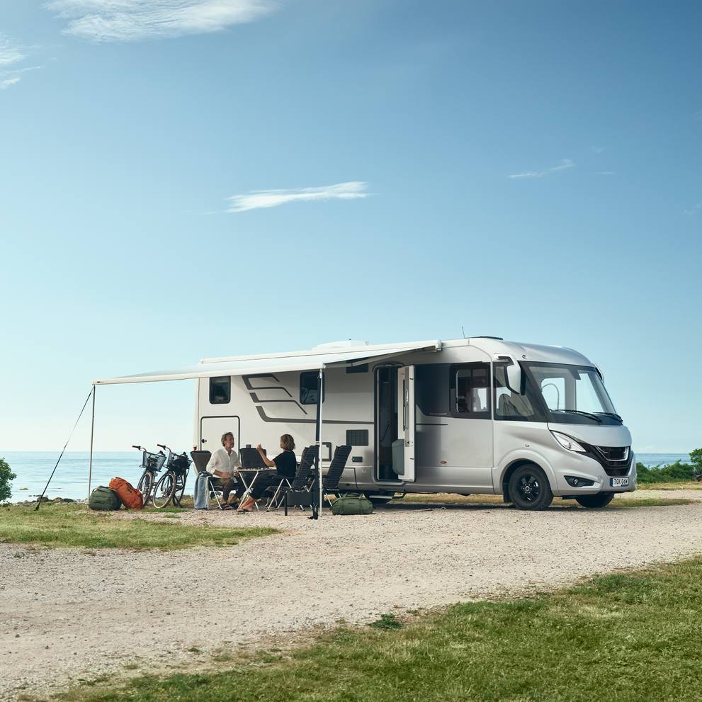 A family sits underneath a Thule Omnistor 8000 motorhome awning on an RV parked near to the water.