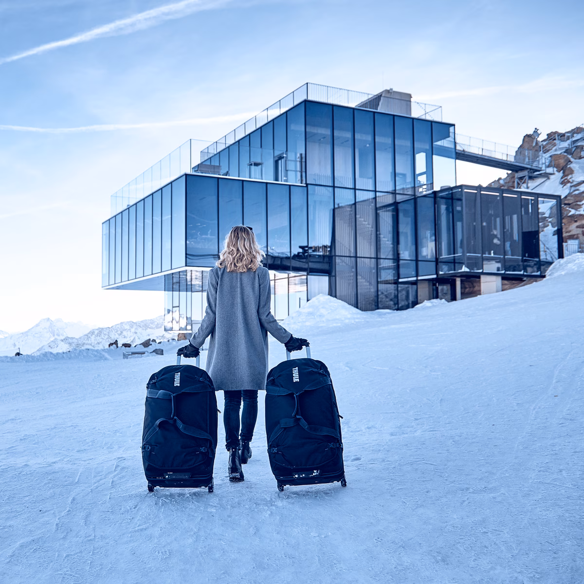In the snow, a woman walks towards a glass building with two Thule Subterra Wheeled Duffel suitcases.