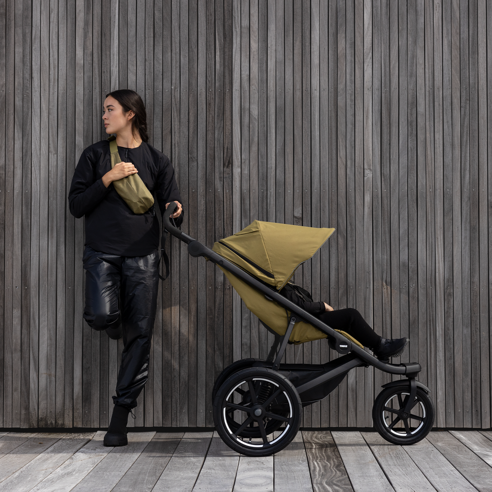 A woman leans against a wooden wall with a green sling bag and a green Thule Urban Glide 3 all-terrain stroller.
