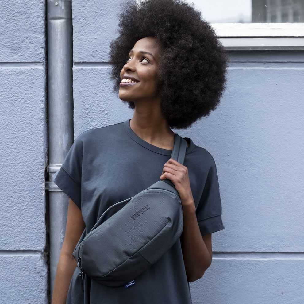 A woman stands against a blue wall and looks up holding a Thule Subterra sling bag.