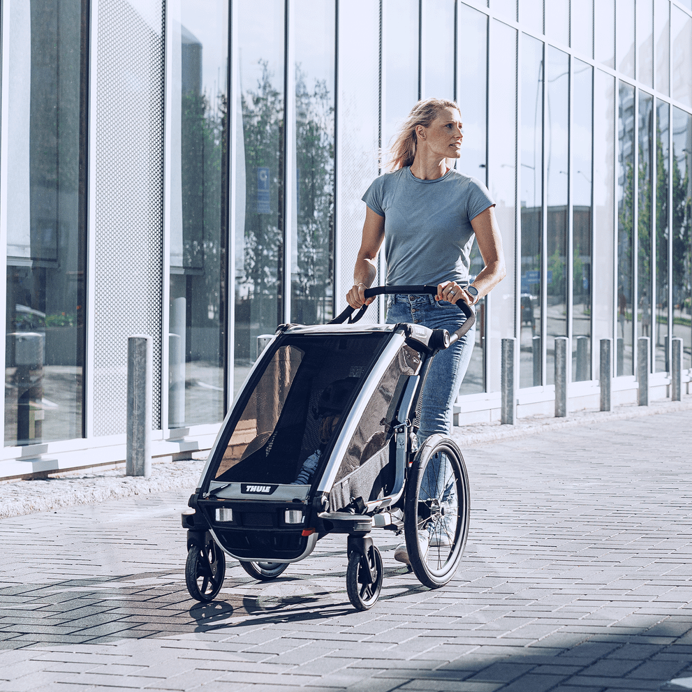 A woman strolls down a city street with her Thule Chariot Lite kids' bike trailer.
