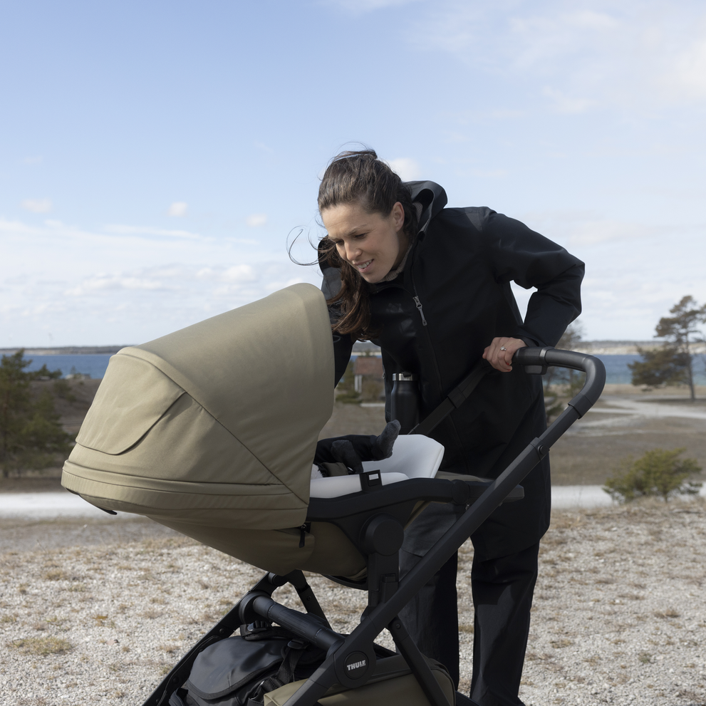 Adult checking on a baby in a Thule Charm stroller bassinet outdoors by the water.