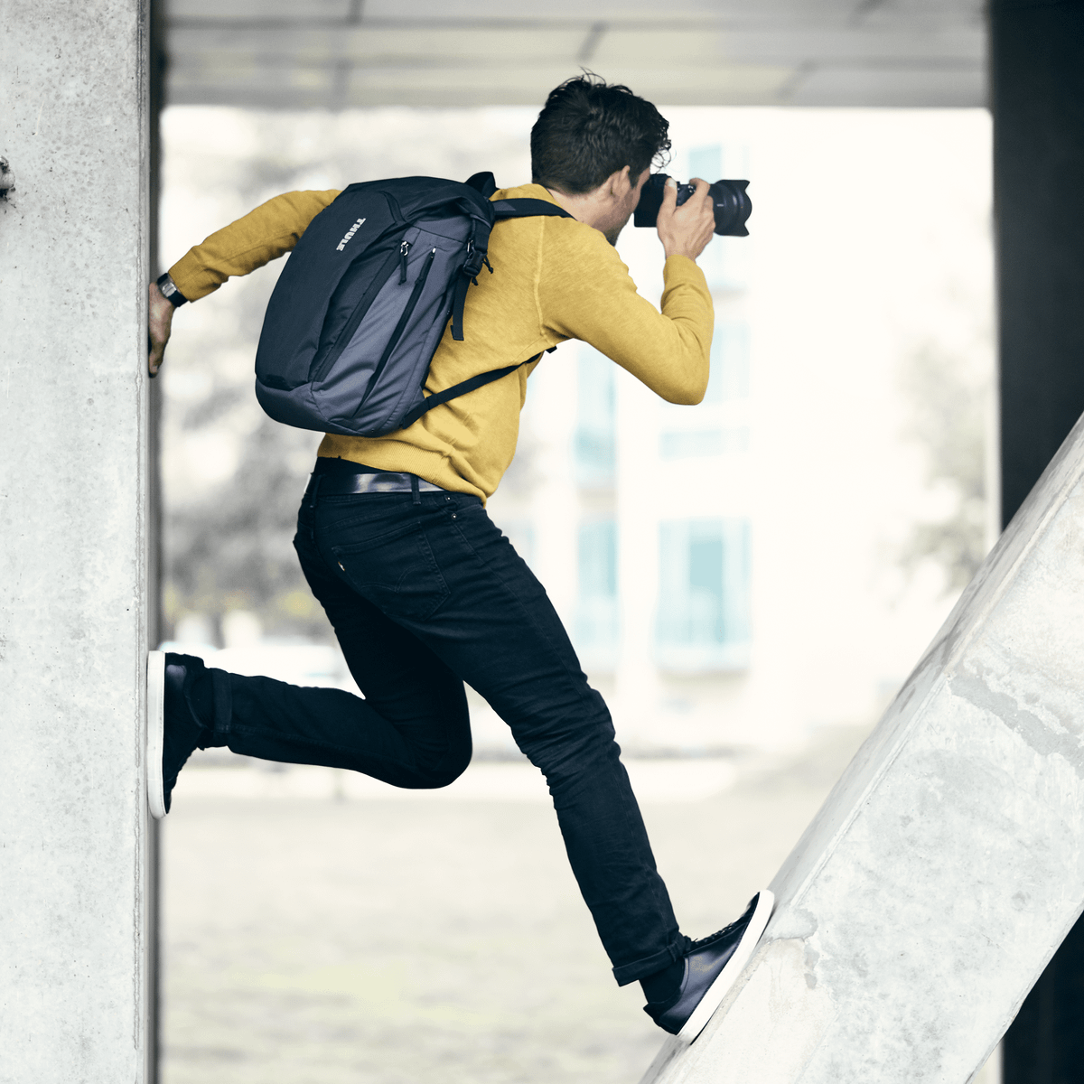 A man stands on a concrete structure taking a picture carrying a blue Thule EnRoute Large DSLR camera backpack.