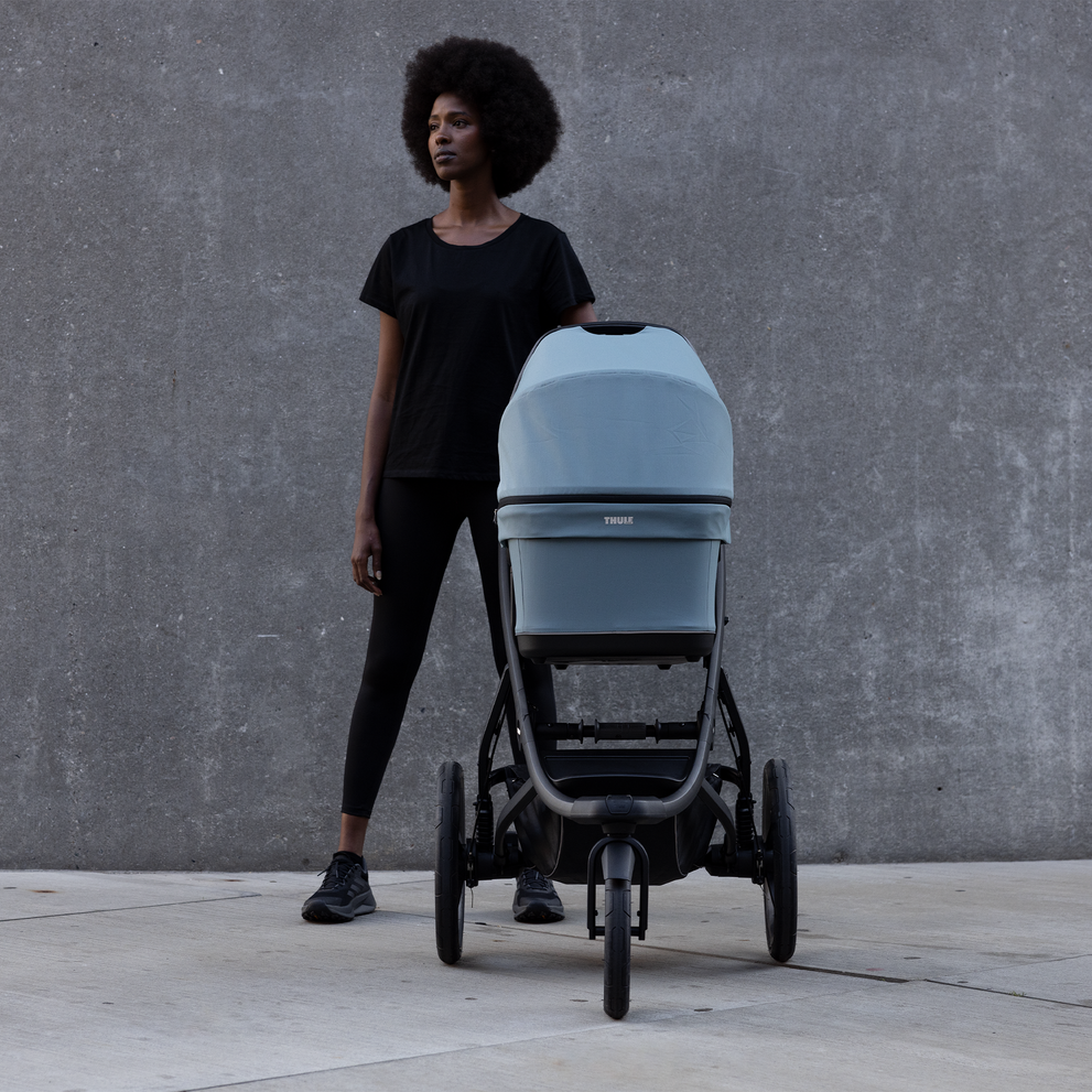 A mother stands next to her blue Thule Urban Glide 3 bassinet stroller against a concrete background.