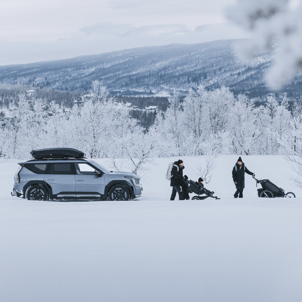 A family is hiking in a snowy landscape, walking with a stroller and a bike trailer away from their car with a Thule Motion 3.
