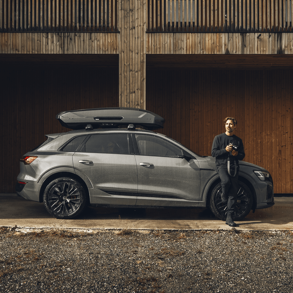 A man leans against a vehicle with a Thule Motion 3 roof box parked next to a wooden wall.