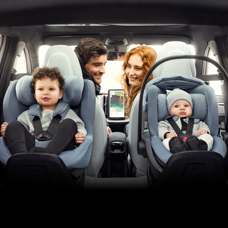 An infant and a toddler is sitting in their car seats in the back of a car while their parents glance back at them.
