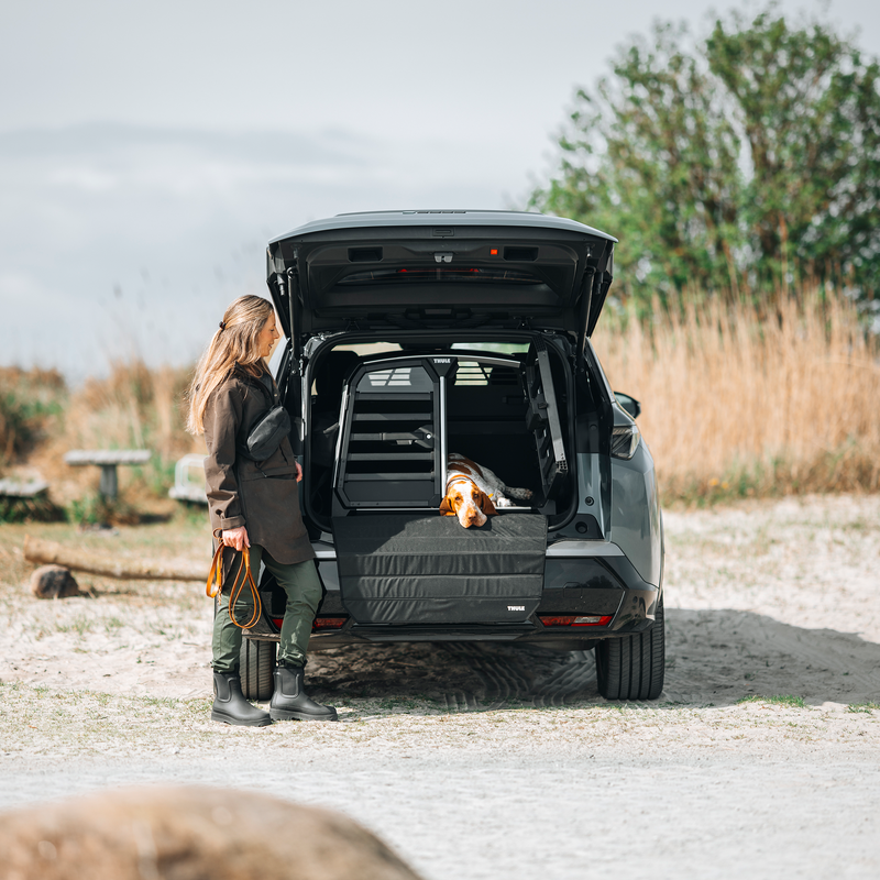 A woman standing next to an open SUV trunk with a dog lying next to the Thule Allax double dog car crate, outdoors on a gravel path.
