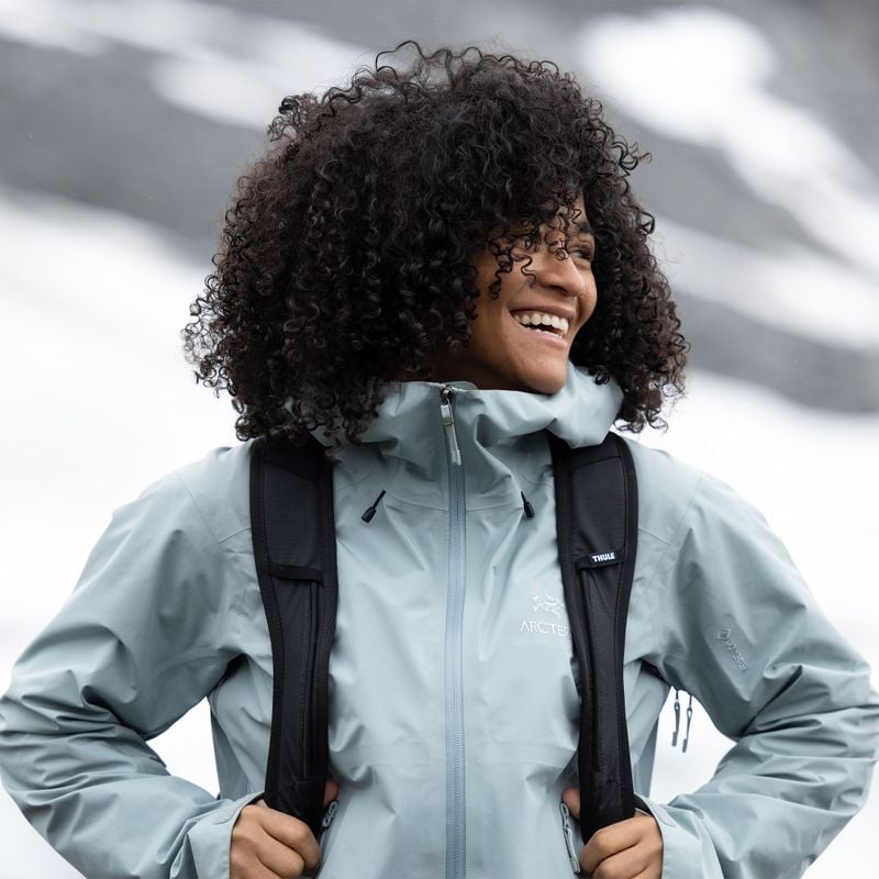 A woman laughs, wearing a blue rain jacket and carrying a black Thule AllTrail hiking backpack.