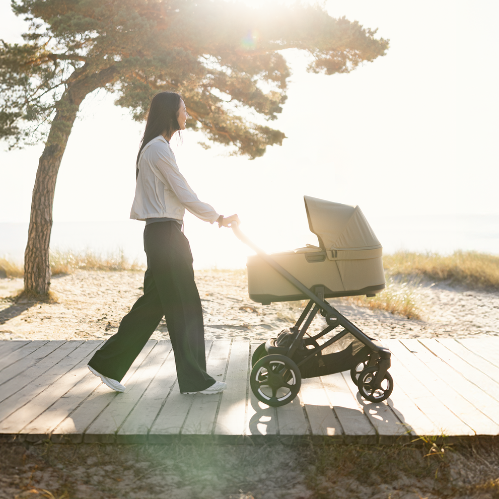 Žena tlačí kočárek Thule Charm po slunné plážové promenádě.