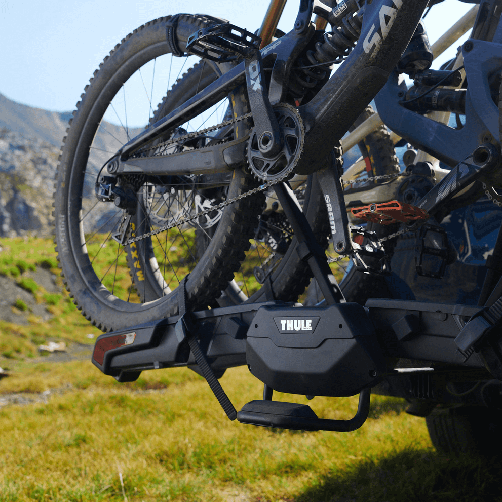 Black bikes are loaded onto a Thule Epos hitch bike rack on a car parked in the grass.