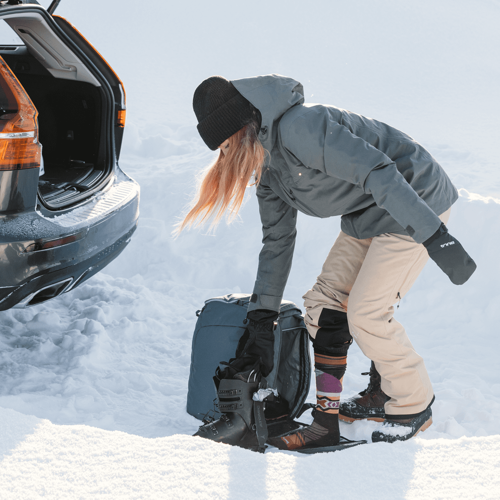 A woman stands in the snow next to the trunk of her car and grabs her 45L Thule RoundTrip Ski Boot Backpack.