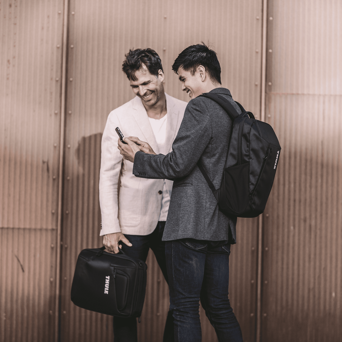 Two men look at the screen of a phone, one is carrying a backpack and the other is holding the Thule Accent Laptop Bag.