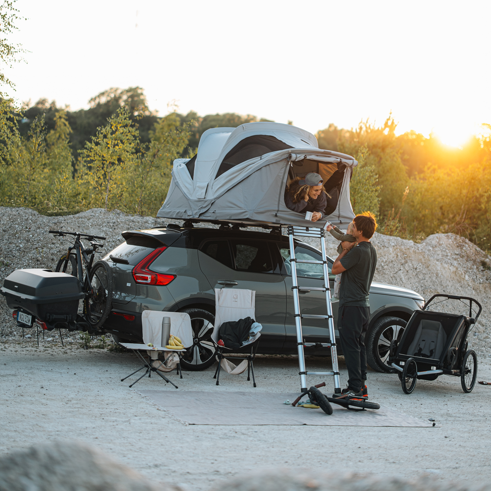 Thule Approach 2 soft-shell rooftop tent unfolded and mounted on the top of a car with one bike and Thule Santu cargo box on the towbar bike rack parked in an outdoor setting and a family interacting by the car.
