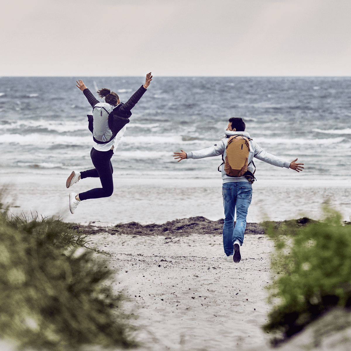 On a cloudy day, two people jump on a beach carrying Thule Stir 25L backpacks.