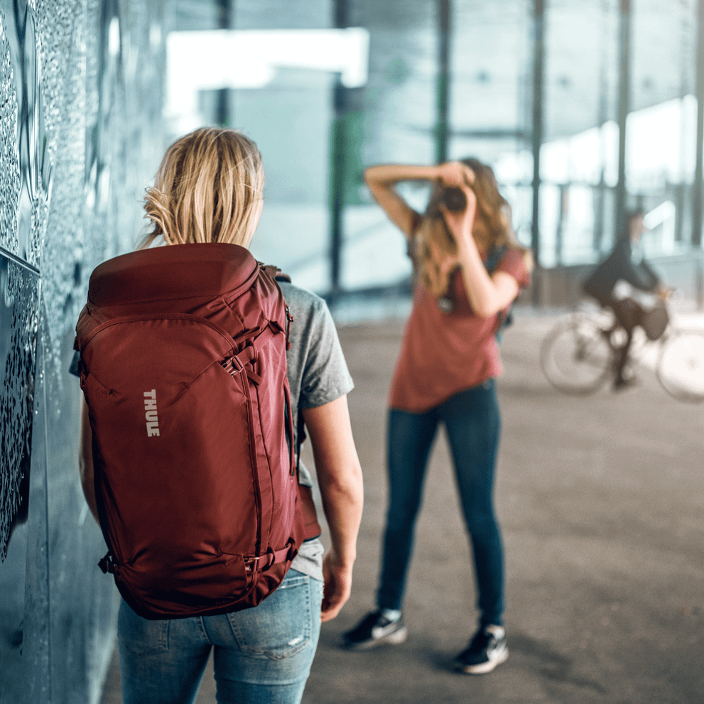 A woman takes a picture of her friend carrying a red Thule Landmark 70L backpack.