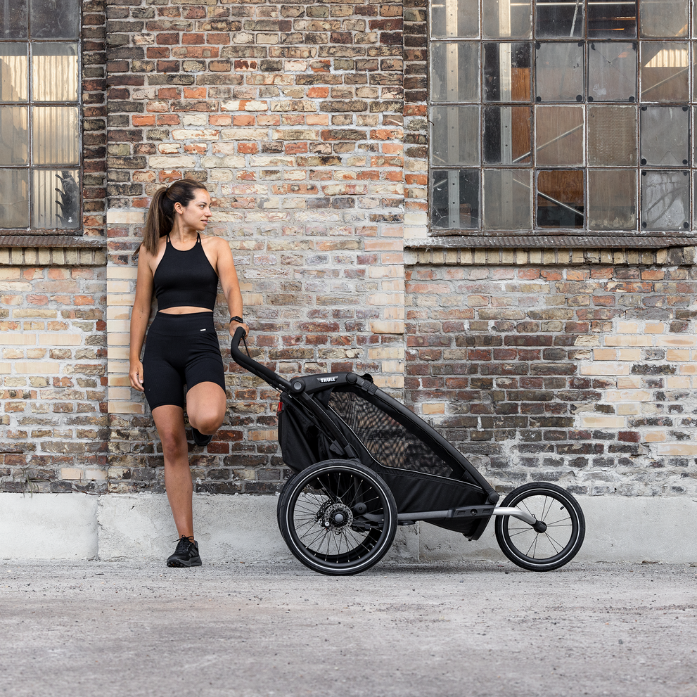 A woman leans against a brick wall with her black Thule Chariot bike trailer.