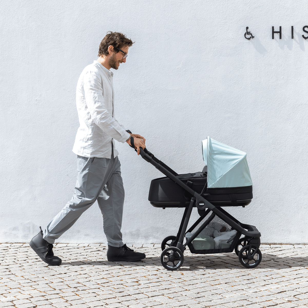 Next to a concrete wall with text, a man walks with his baby in a blue Thule Shine baby stroller.