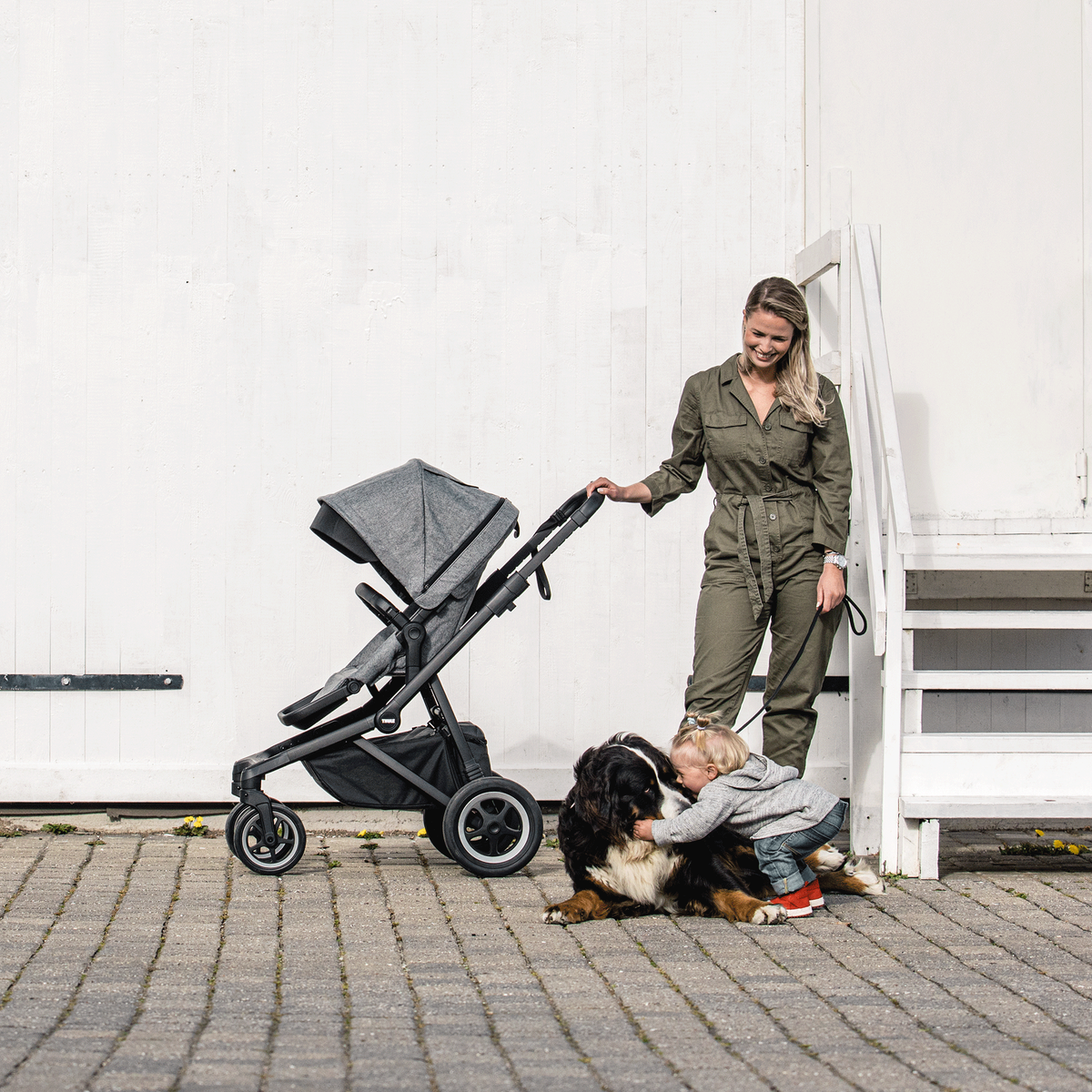 Next to a white garage door, a woman looks at her child while pushing a gray Thule Sleek baby stroller.