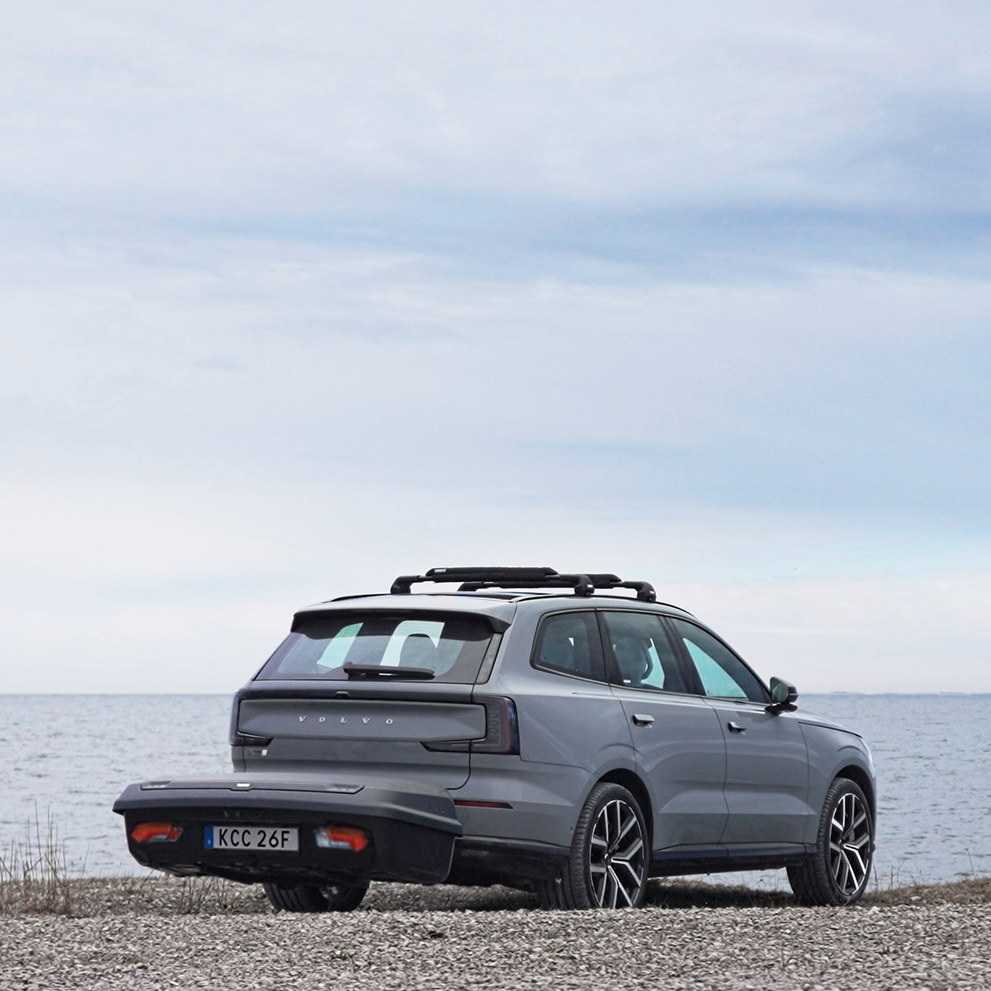 A dark grey car with Thule Arcos XL hard-shell rear cargo carrier mounted on the back, is parked by a shoreline, with roof racks on the top of the car.
