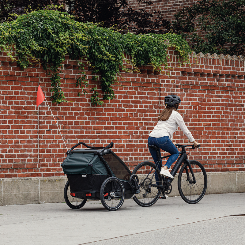 A woman cycling through a city with a Thule Chariot Cab attached to the back of her bike.