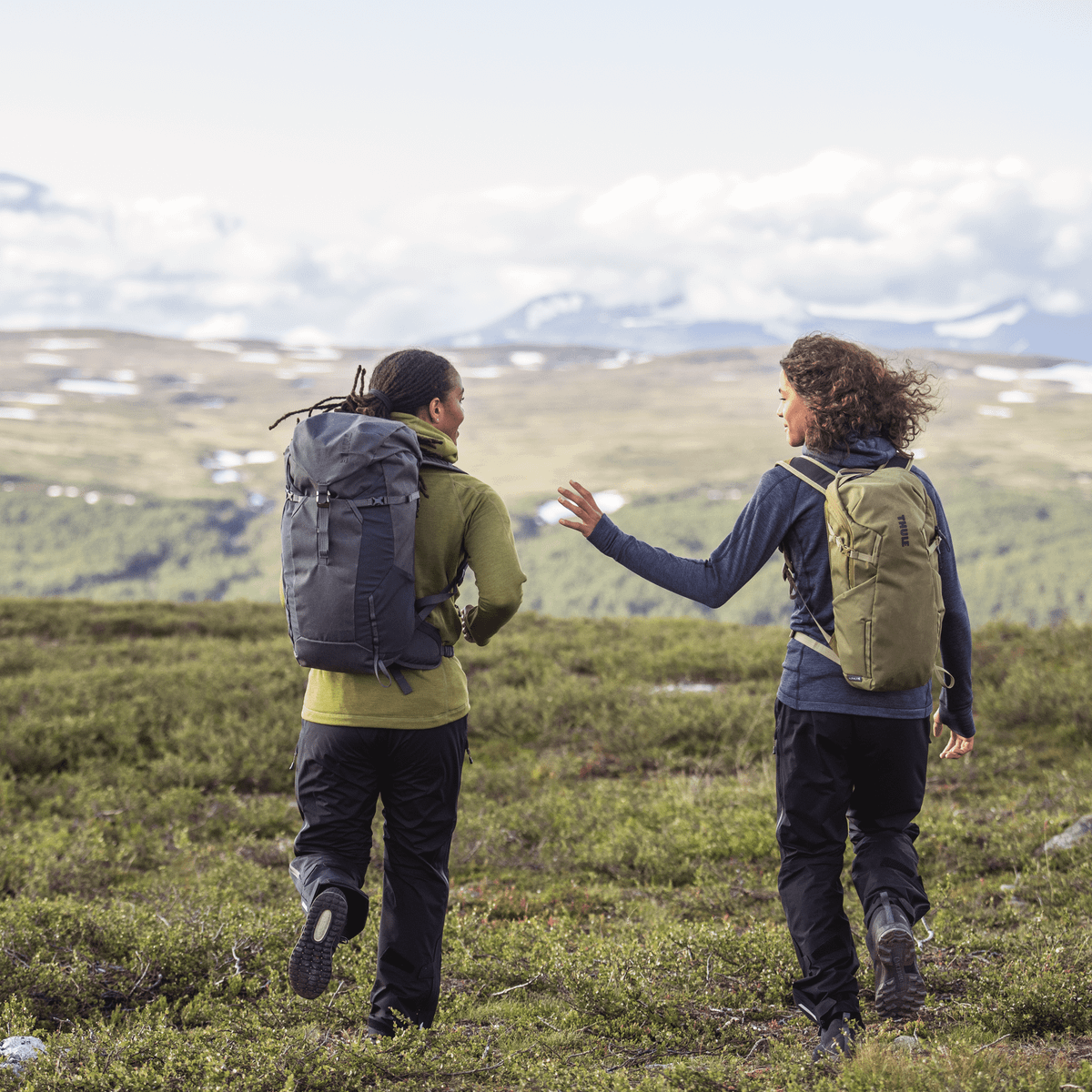 Two people walk through a field carrying green and black Thule AllTrail X hiking backpacks.