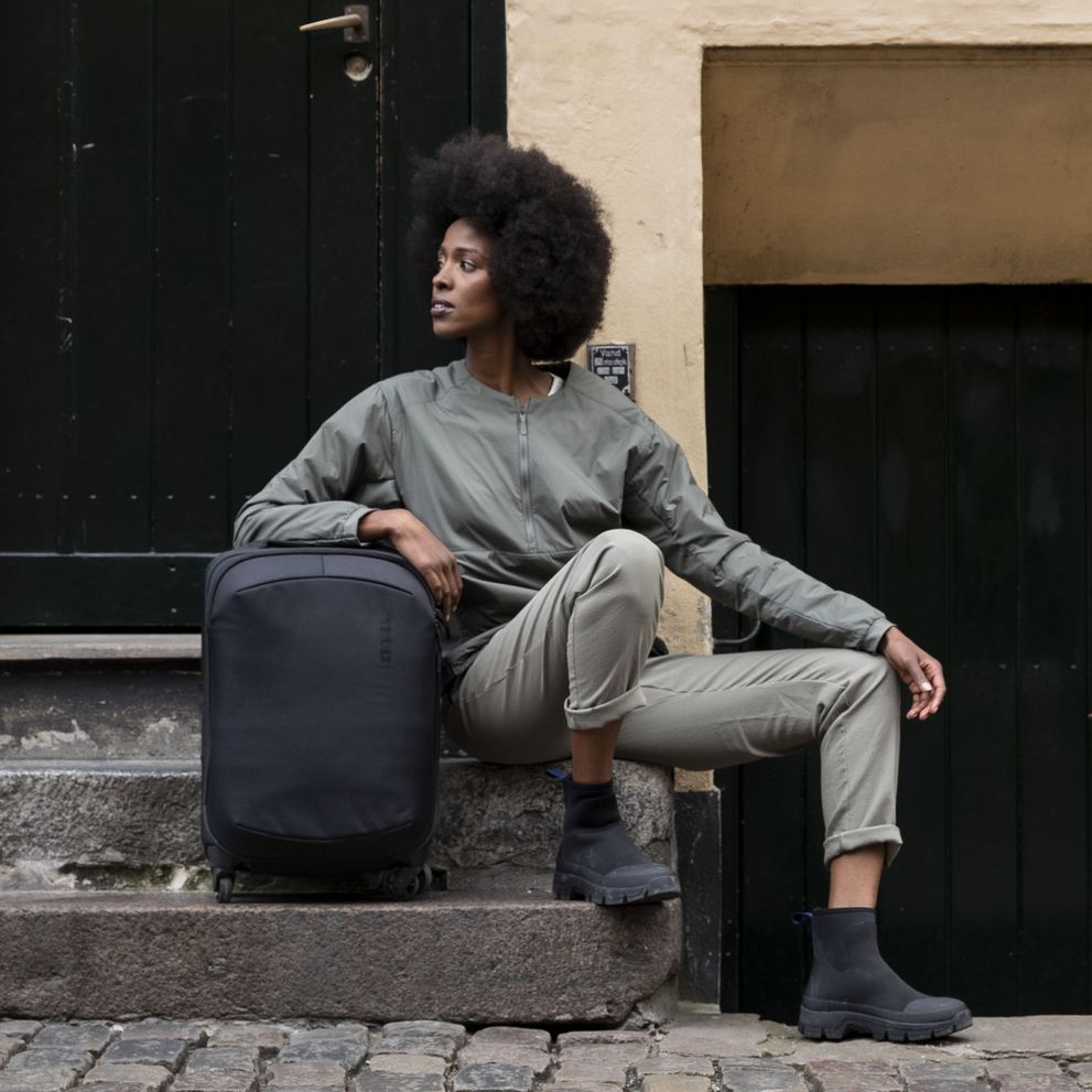 A woman sits on a stoop holding a black Thule subterra carry-on suitcase.