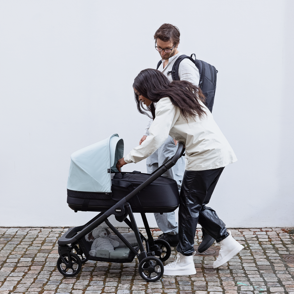 Parents tend to their baby sleeping inside a blue Thule Shine bassinet.