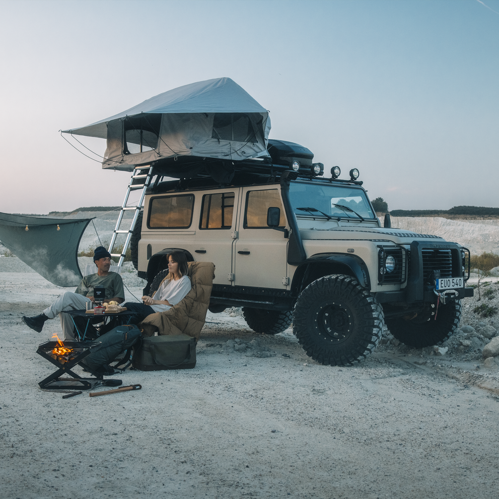 Thule Foothill 2 soft-shell rooftop tent unfolded and mounted on top of a car with a roofbox, parked in an outdoor setting with a man and a woman sitting by a camp fire.