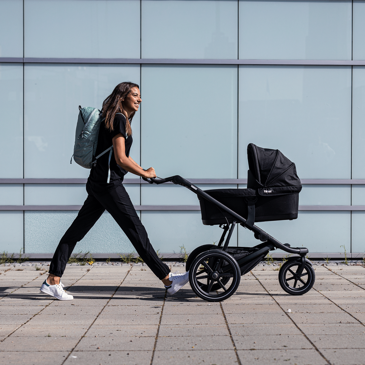 A woman walks down a city street with the black Thule Urban Glide 2 jogging stroller and bassinet.