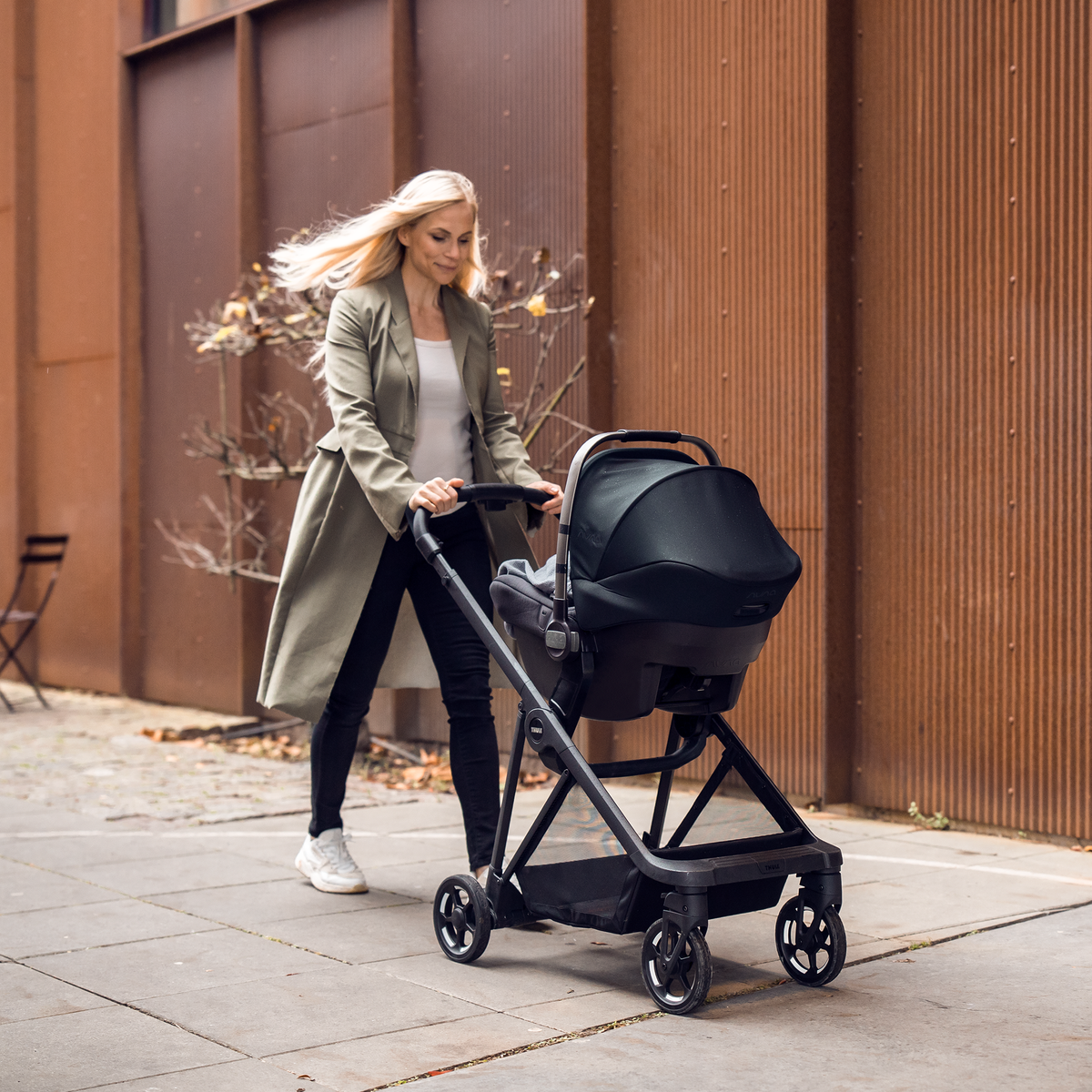 A woman walks down a street with her baby in a stroller with a car seat using the Thule Shine Car Seat Adapter.
