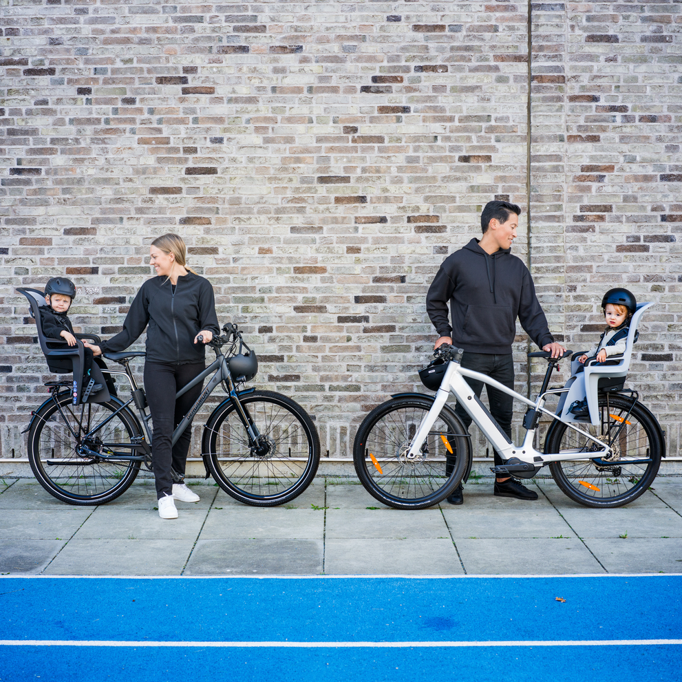 Two parents stand against a brick wall with bikes and their kids in Thule RideAlong2 child bike seats.