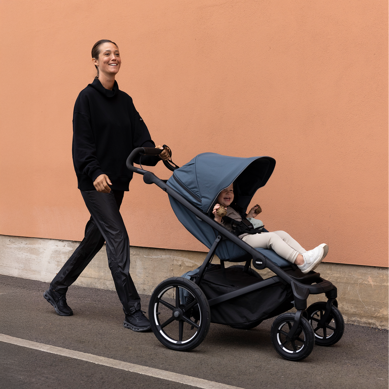 Donna che spinge passeggino Thule Urban Glide a 4 ruote con seggiolino auto vicino a muro di cemento.