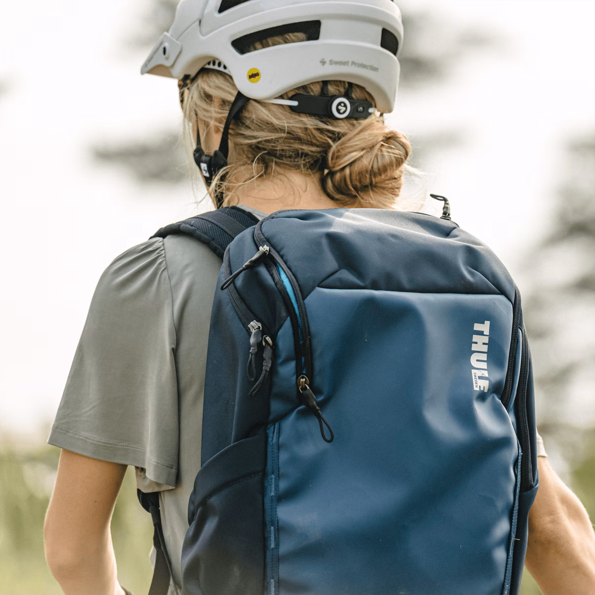 A woman with a white helmet walks with a blue Thule Chasm backpack.