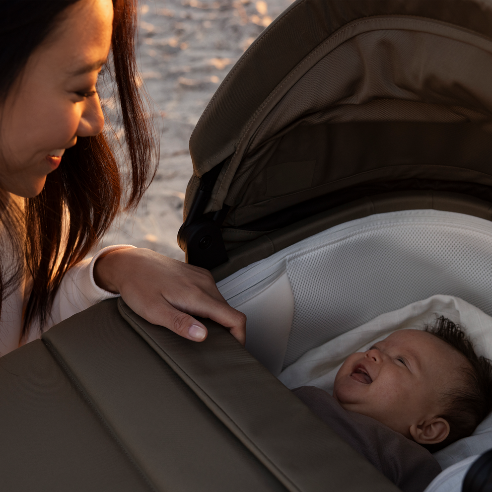 Usmívající se dospělý se dívá na spokojené miminko ležící v Thule Charm bassinet na písečné pláži v teplém slunečním světle.