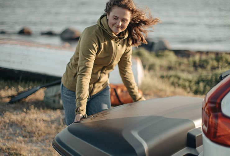 A woman attaching a Thule Arcos cargo box on a car that is parked next to the ocean