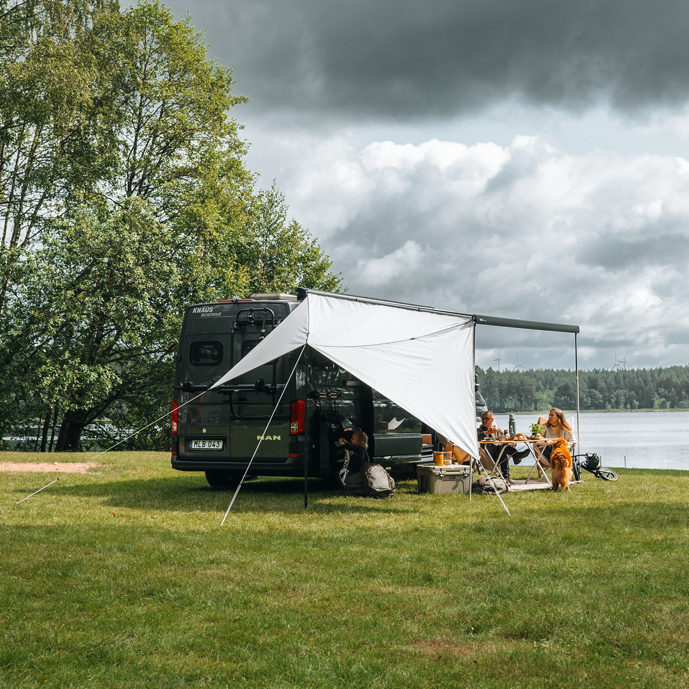 Camper van with Thule awning and side panel, set up by a lake with people and a dog enjoying a meal outdoors.