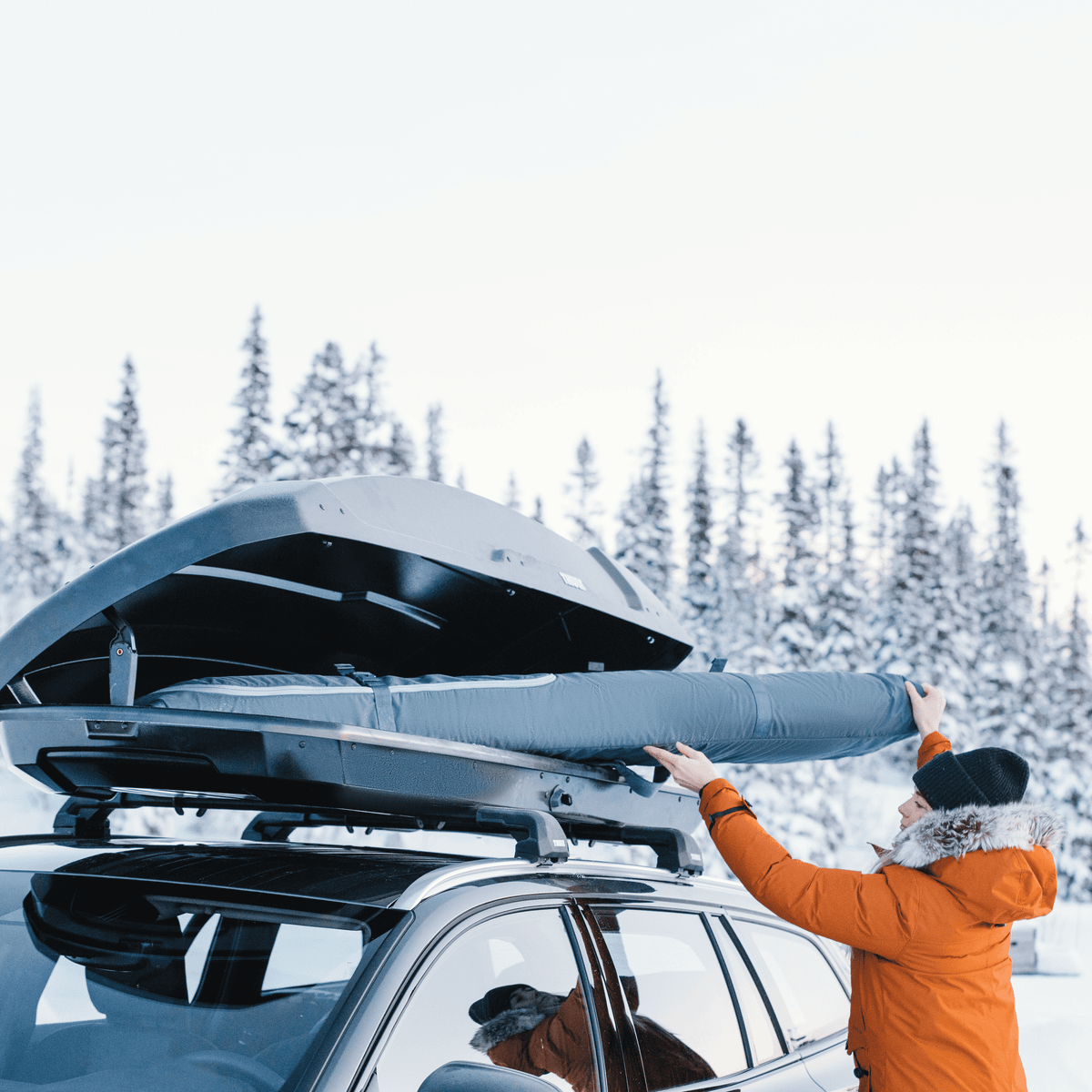 A person in the snow loads a Thule RoundTrip Snowboard Bag into the roof box of their car.